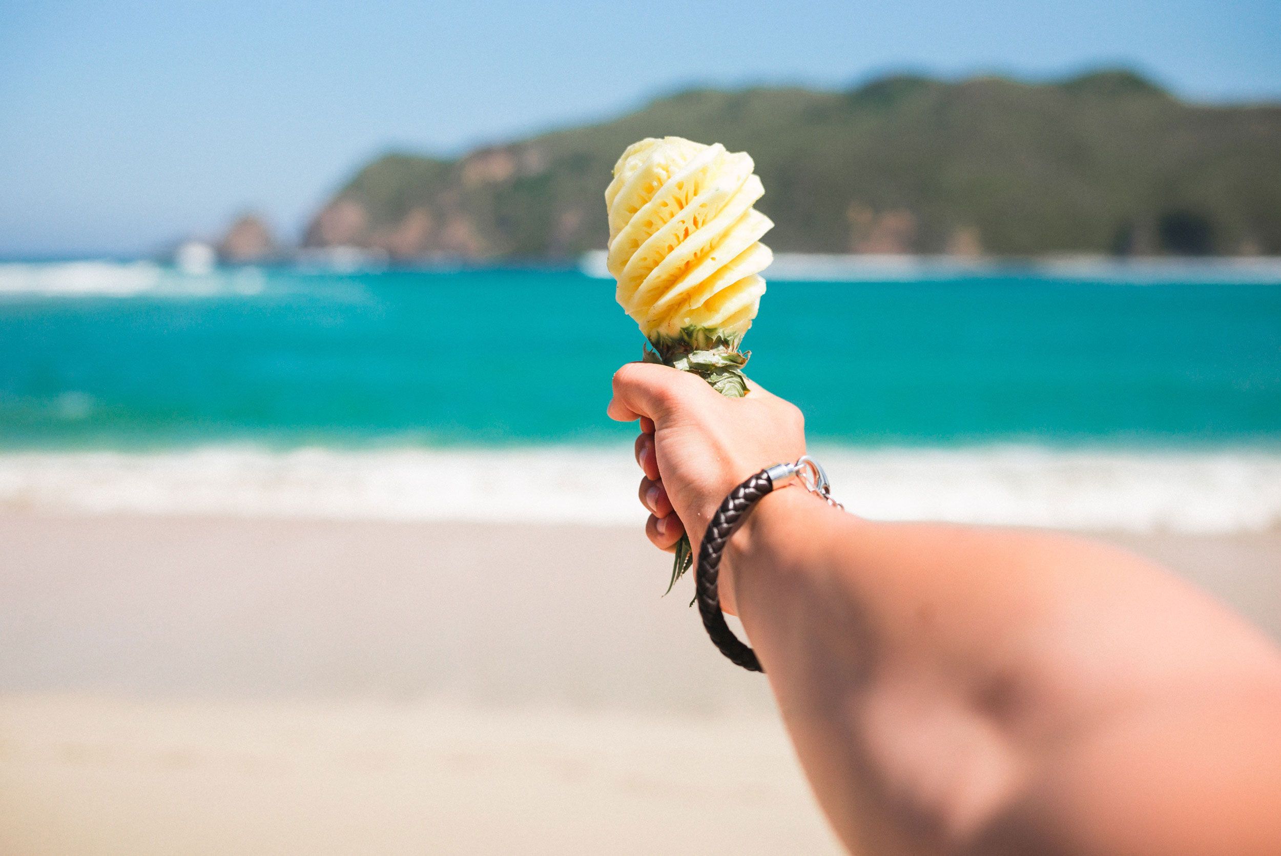 Ein ausgestreckter Arm hält eine Ananas, die wie ein Mikrophon geschnitzt ist, in Richtung Strand